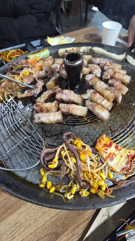 솥뚜껑에 가득 찬 삼겹살, 김치, 콩나물, 고사리