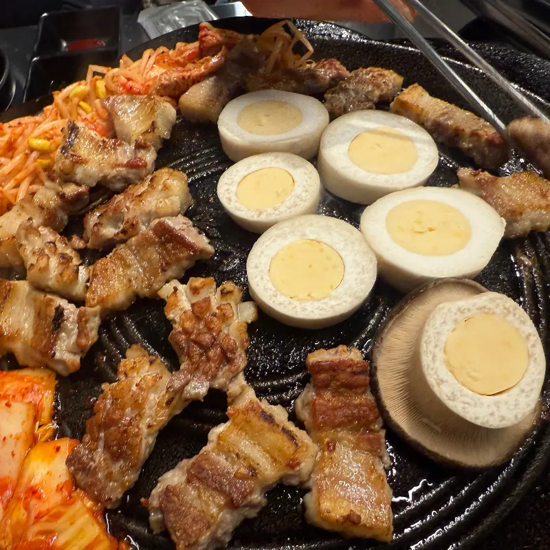 솥뚜껑에 구워진 삼겹살, 김치, 콩나물, 계란