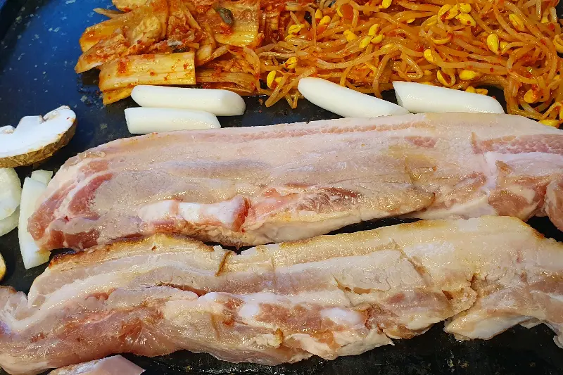 불판 위에 올려진 삼겹살, 김치, 콩나물무침의 조화