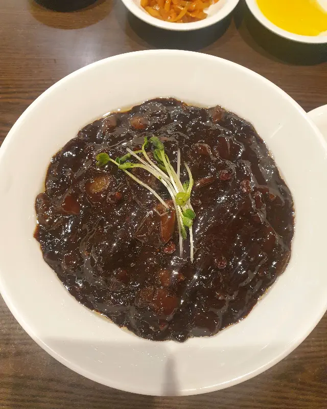 검은 짜장 소스가 면 위에 덮여있는 짜장면이 흰색 그릇에 담겨 테이블 위에 놓여 있다.