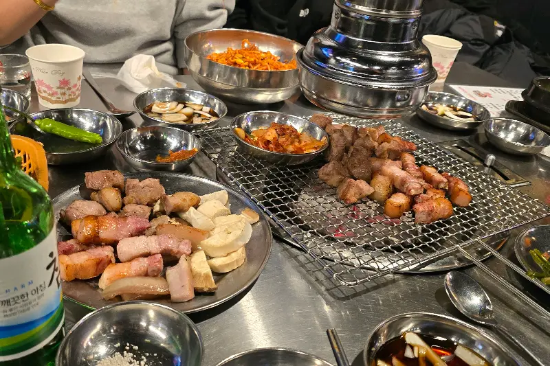 푸짐하게 차려진 테이블, 석쇠 위에는 초벌된 삼겹살이 놓여 있다.