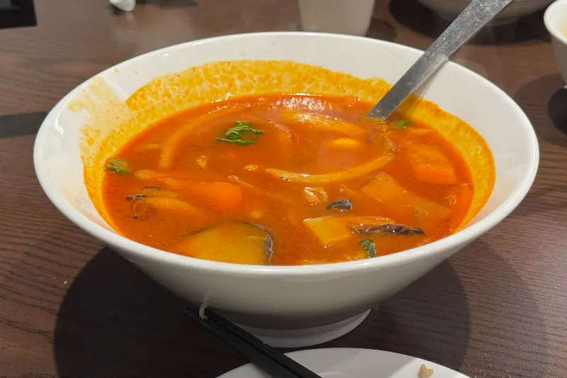 하얀색 짬뽕 그릇에 담긴 붉은색 국물의 짬뽕이 테이블 위에 놓여 있다. 스푼이 짬뽕 안에 담겨 있다.