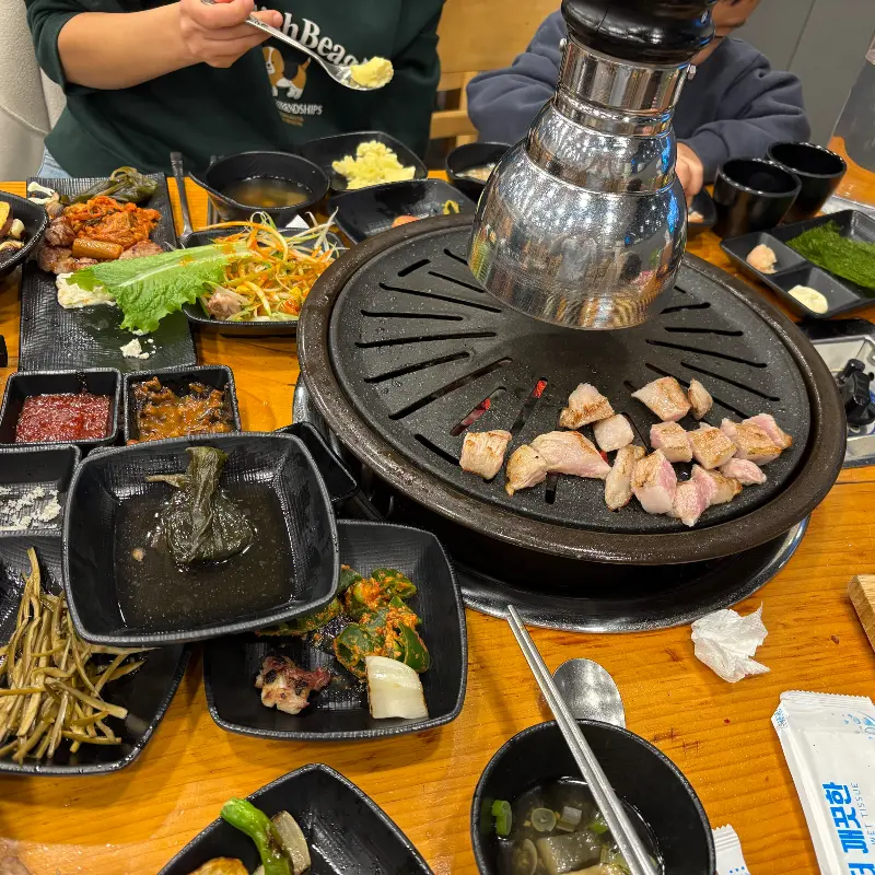 숯불 테이블에 오겹살, 밑반찬, 냉면 등이 가득 차려진 푸짐한 한 상 차림 사진이다.