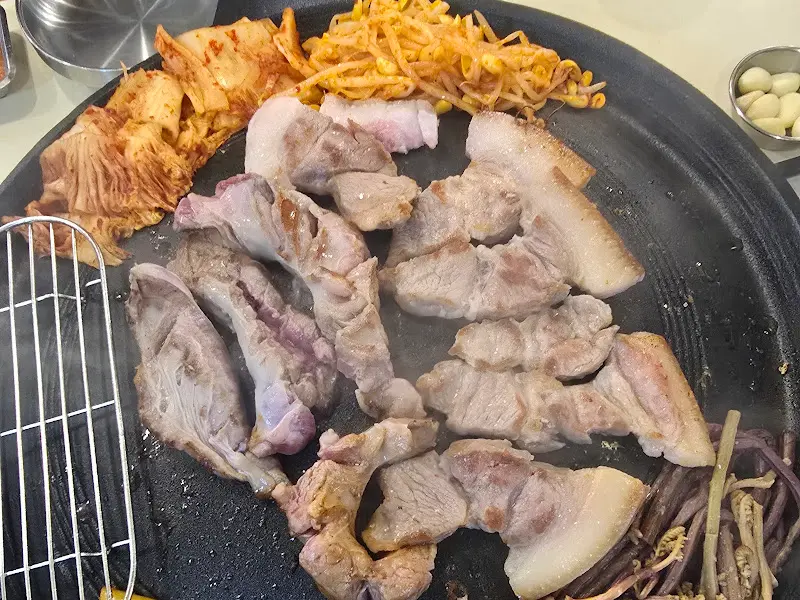불판 위에 올려진 삼겹살, 김치, 콩나물, 고사리