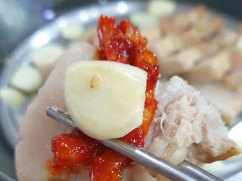 보쌈을 젓가락으로 집어 올린 모습, 보쌈 위에 붉은 양념과 마늘 한쪽이 올려져 있다.