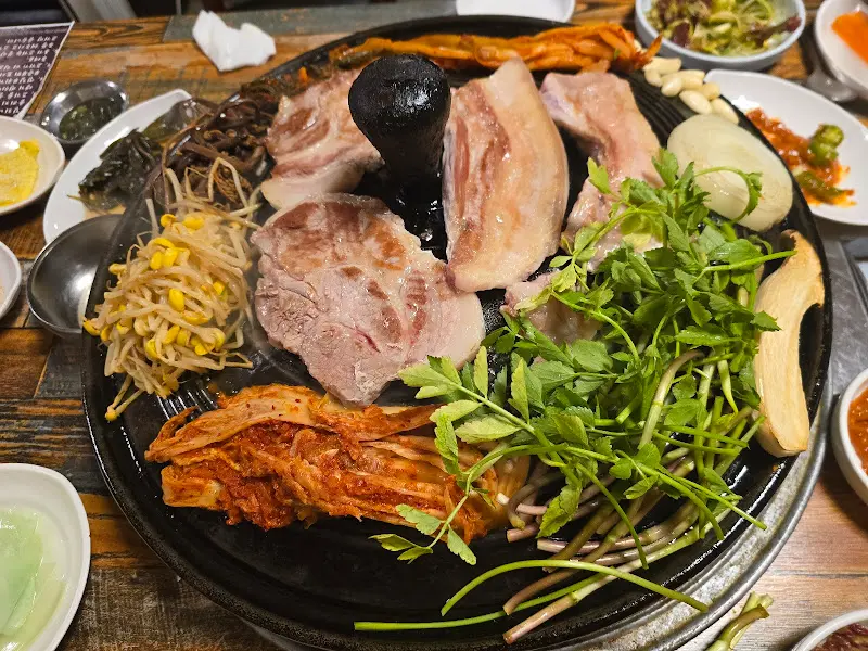 솥뚜껑 위에 삼겹살, 김치, 콩나물, 고사리가 푸짐하게 올려져 있는 모습