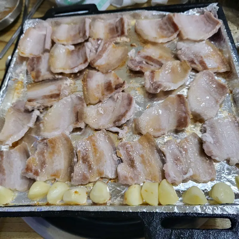 불판 위에서 노릇노릇하게 익어가는 삼겹살, 마늘도 함께 구워 풍미를 더한다.