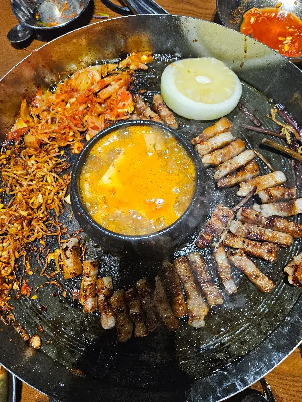 솥뚜껑 위에 구워진 삼겹살, 김치, 콩나물, 그리고 된장찌개의 조화
