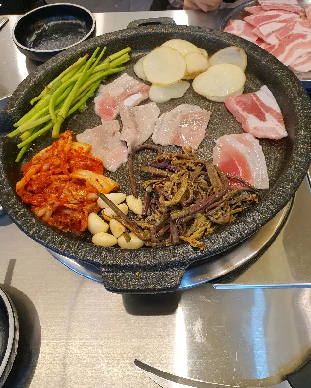 불판 위에 냉동 삼겹살과 김치, 콩나물, 미나리, 감자가 함께 구워지고 있는 모습
