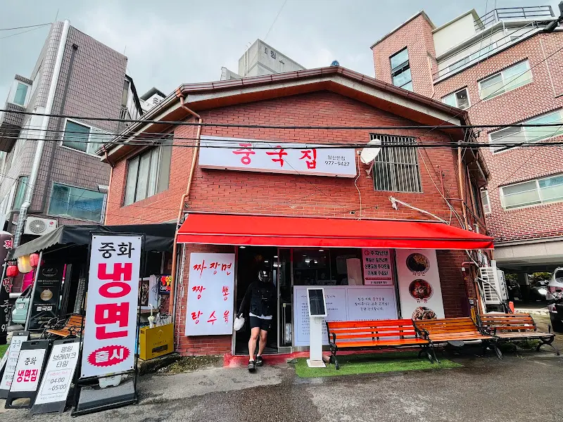 붉은 벽돌 건물에 '중국집'이라고 적힌 간판이 달려 있다. 빨간색 천막이 쳐져 있고, 그 아래에 테이블과 의자가 놓여 있다.