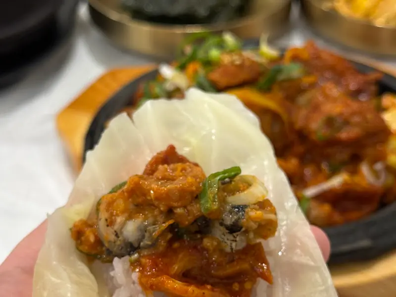쌈 위에 밥, 제육볶음, 어리굴젓을 올려 한 입 가득 먹는 모습