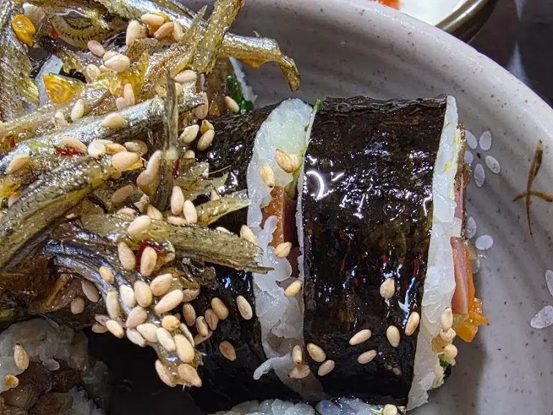 테이블에 놓인 멸치 김밥의 단면을 확대한 사진. 김밥 속 재료와 멸치가 조화롭게 어우러져 있고, 깨소금이 뿌려져 있어 더욱 먹음직스럽다.