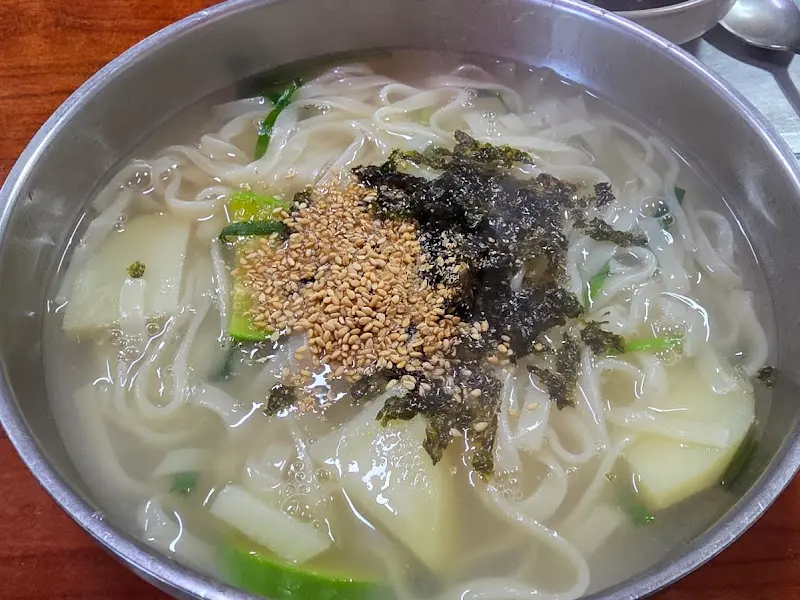 스테인리스 그릇에 담긴 칼국수 사진. 면발이 탱글탱글하고, 김가루와 깨소금이 듬뿍 뿌려져 있어 먹음직스럽다.