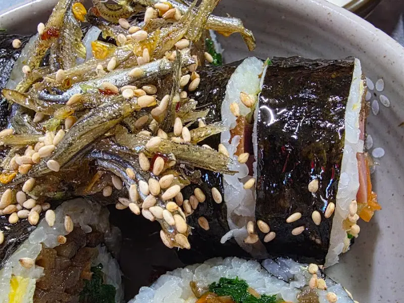 멸치 김밥 위에 멸치가 수북이 쌓여 있는 모습. 멸치와 김밥의 조화가 독특하고 맛있어 보인다.