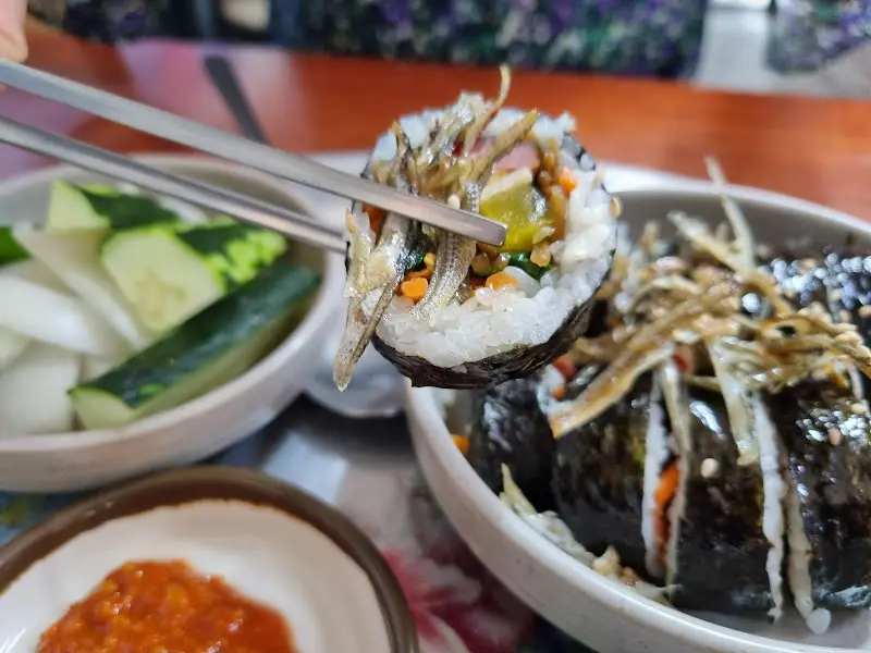 젓가락으로 멸치 김밥을 집어 올린 모습. 멸치가 듬뿍 올려져 있어 먹음직스럽다.