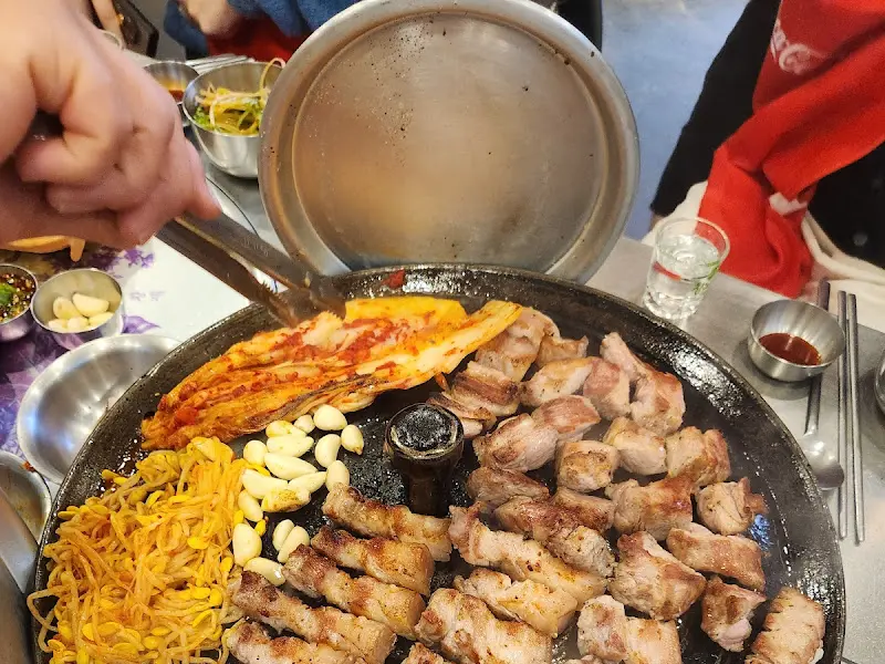 불판 위에서 맛있게 익어가는 삼겹살과 김치, 콩나물, 마늘