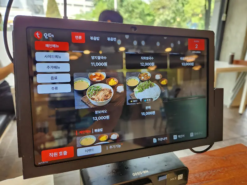 테이블 위에 놓인 태블릿 메뉴판. 다양한 메뉴 사진과 가격 정보가 표시되어 있다.