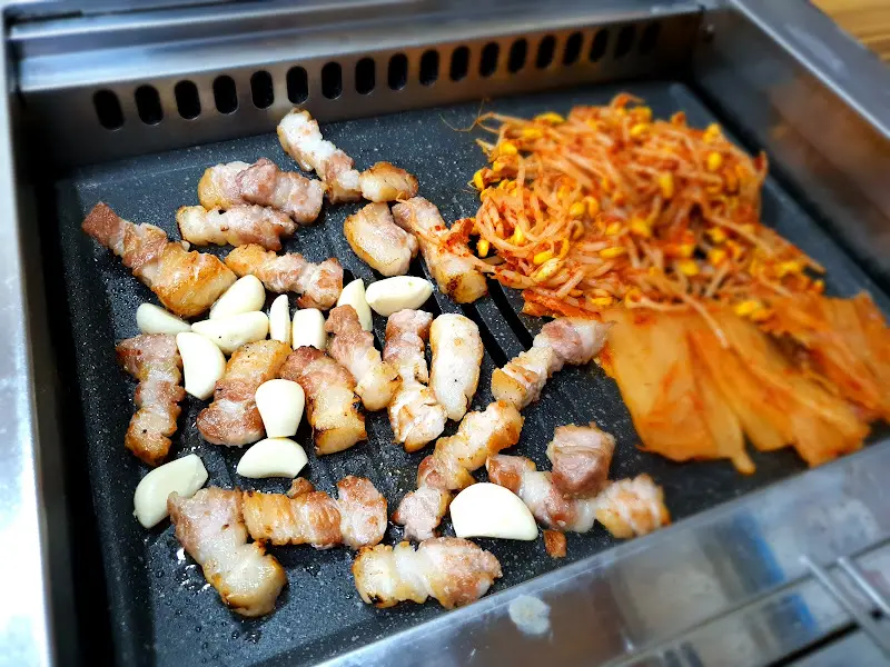 불판 위에 구워진 삼겹살, 김치, 마늘, 콩나물