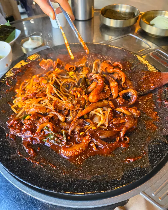 철판 위에서 쭈꾸미와 콩나물이 매콤한 양념에 볶아지고 있는 모습