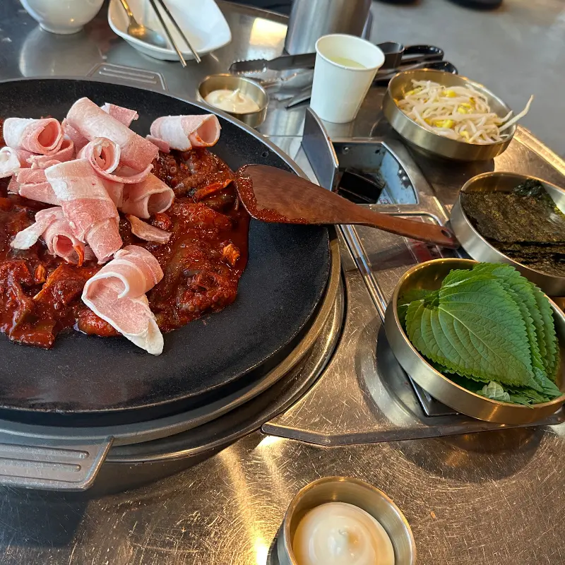 쭈꾸미 철판 위에 대패 삼겹살이 얹어져 있고, 깻잎, 김, 콩나물 등의 다양한 쌈 재료가 놓인 테이블 전경