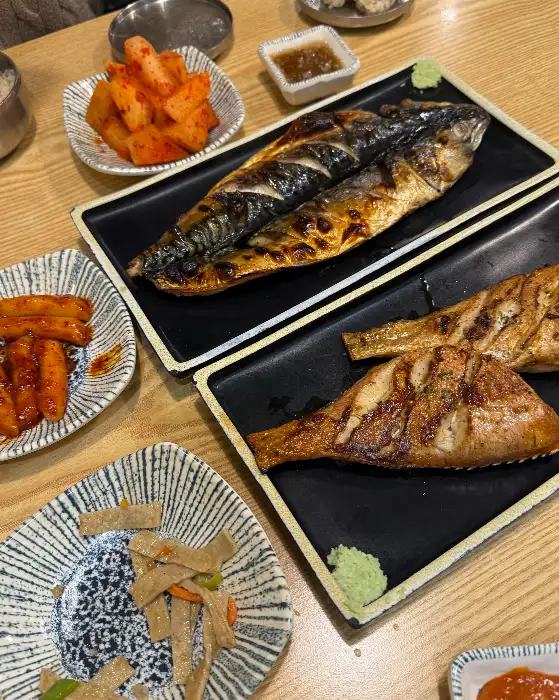 테이블 위에 놓인 고등어구이와 떡볶이, 그리고 다른 밑반찬들의 모습. 먹음직스러운 생선구이가 메인 요리임을 보여준다.