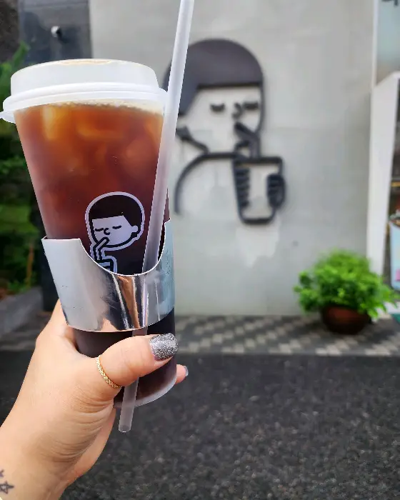 손으로 들고 있는 아이스 아메리카노 컵과 뒤로 보이는 카페 로고