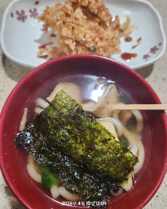 우동 그릇에 김과 어묵 등이 들어 있고, 뒤쪽에는 튀김 요리가 담긴 접시가 놓여 있는 모습