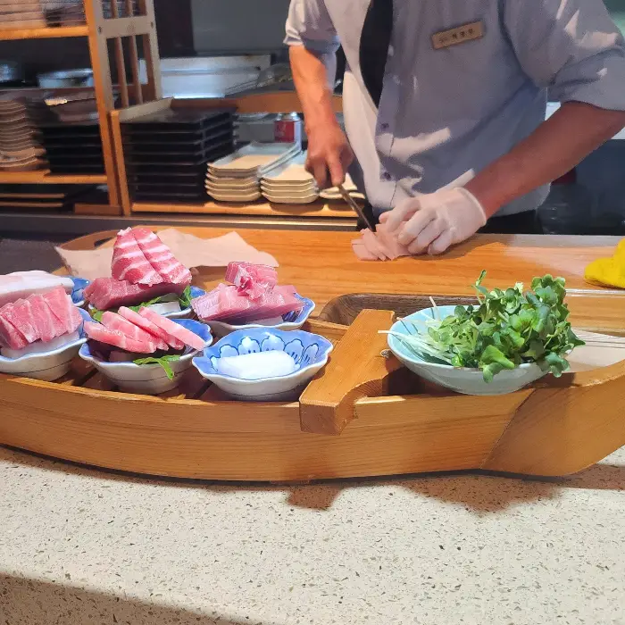 요리사가 나무 도마 위에서 초밥을 만들고 있는 모습, 앞에 신선한 회와 채소가 놓여 있다.