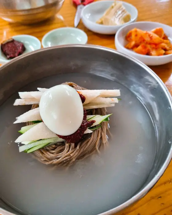 살얼음이 동동 뜬 맑은 육수의 물막국수. 삶은 달걀, 오이채, 김가루, 그리고 빨간 양념장이 조화롭게 올라가 있습니다.
