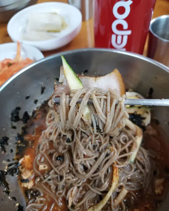 젓가락에 집힌 비빔막국수 면발. 빨간 양념이 듬뿍 묻어 있고, 삶은 달걀 고명과 오이채가 보입니다.