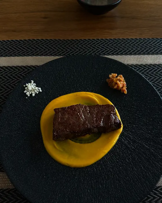 블랙 접시 위에 노란색 퓨레가 깔리고 그 위에 두툼한 고기가 올라간 요리. 곁들임으로 흰색 가루와 붉은색 다진 재료가 보인다.