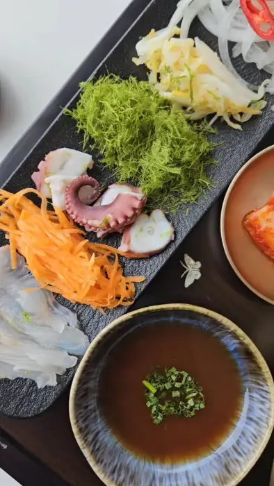 접시 위에 채 썰어진 오이, 당근, 그리고 문어 다리 조각들이 담겨 있고, 그 위로 파릇한 채소가 흩뿌려져 있다. 접시 가장자리에는 소스가 담긴 그릇이 보인다.