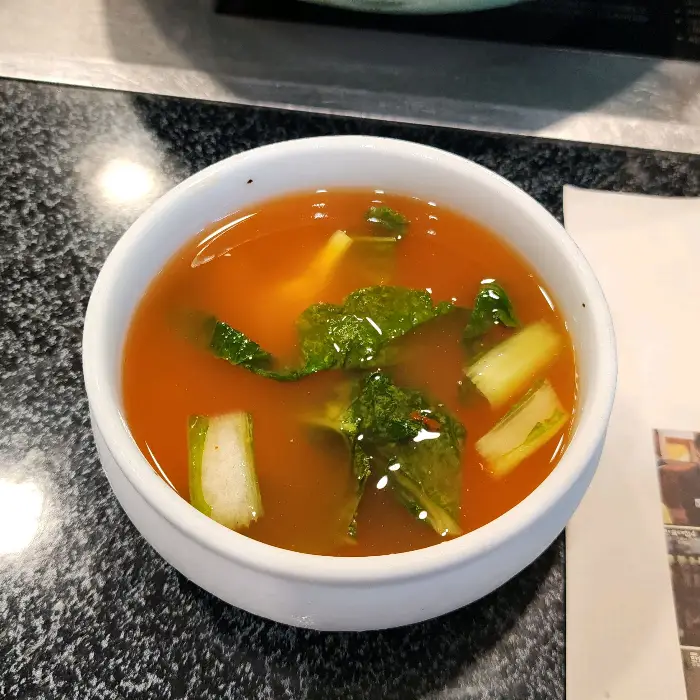 흰색 그릇에 담긴 붉은 국물에 녹색 채소와 흰색의 썰린 채소가 들어 있다. 맑은 국물 위로 잎채소 조각들이 떠 있다.