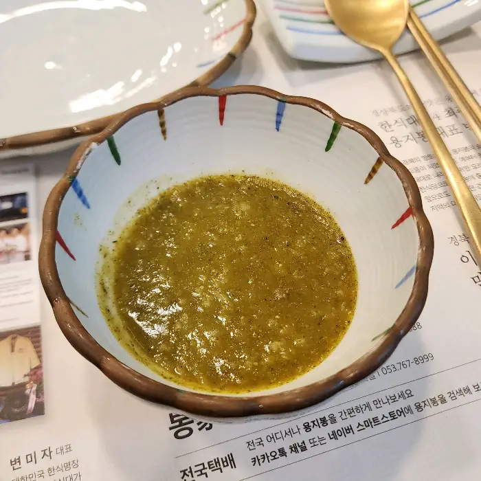 독특한 모양의 흰색 그릇에 담긴 연한 녹색의 소스가 있다. 소스 위에는 작은 녹색 알갱이들이 뿌려져 있다.