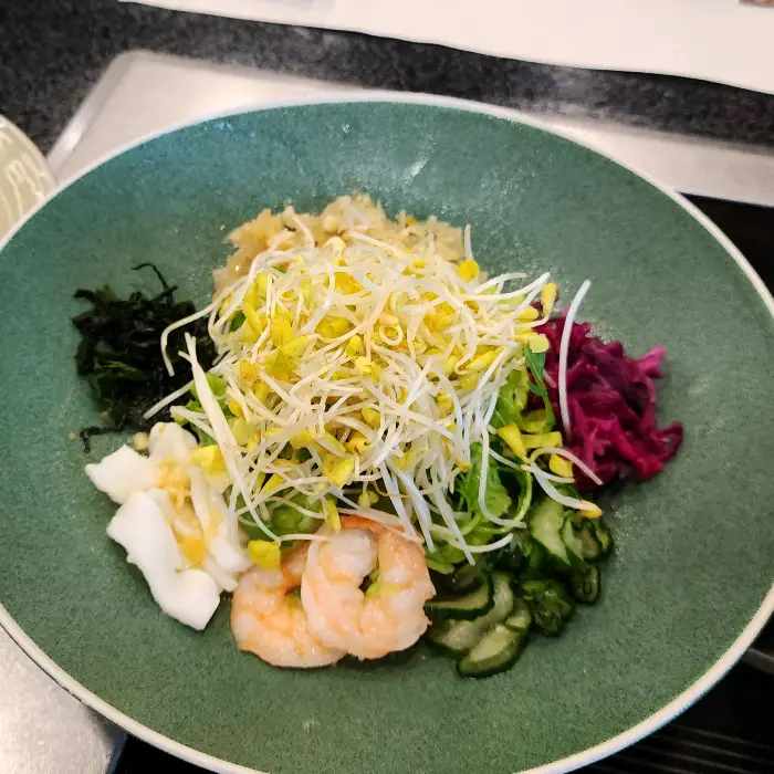 넓은 녹색 접시에 다양한 채소와 새우, 해초류가 담겨 있다. 새싹 채소와 붉은색, 보라색 채소, 오이 조각, 그리고 두 마리의 분홍색 새우가 돋보인다.