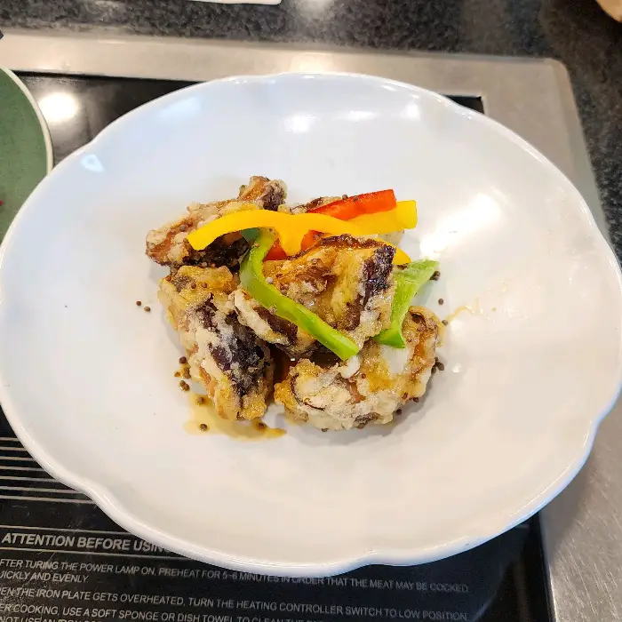 흰색 접시에 튀김 옷을 입혀 튀겨진 재료 조각들이 담겨 있다. 그 위로 얇게 썰린 피망, 파프리카, 노란색 채소가 얹혀 있다.