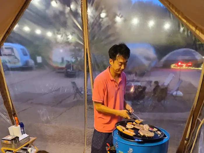 한 남자가 텐트 안에서 바베큐 그릴에 고기를 굽고 있는 모습입니다. 텐트 너머로 캠핑장의 풍경이 보입니다.