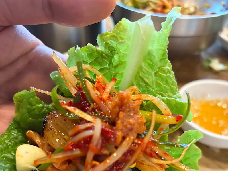 흑돼지, 콩나물무침, 마늘, 멜젓을 쌈 채소에 싸서 한 입