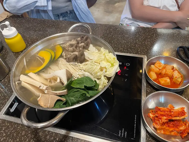 만두전골 냄비 안에 만두, 채소, 버섯 등이 푸짐하게 담겨 있고, 깍두기와 김치가 곁들여져 있는 모습