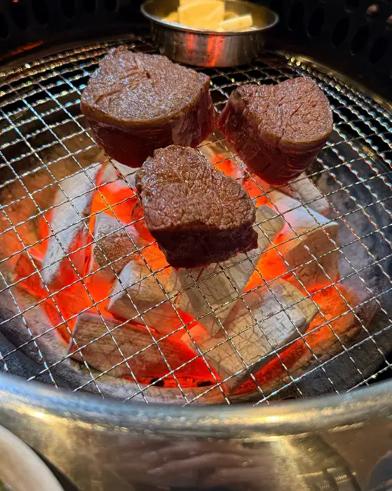 숯불 위에서 잘 익고 있는 두툼한 스테이크 조각. 겉면은 먹음직스럽게 익었고, 속은 촉촉해 보인다.