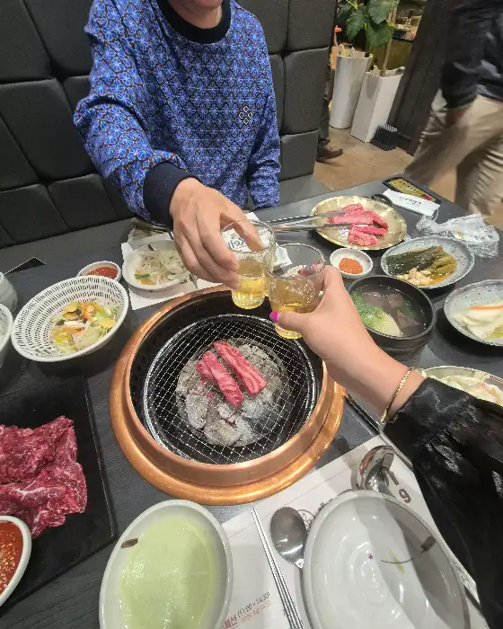 두 사람이 술잔을 부딪치며 건배하는 모습, 테이블에는 고기와 반찬이 놓여 있다.