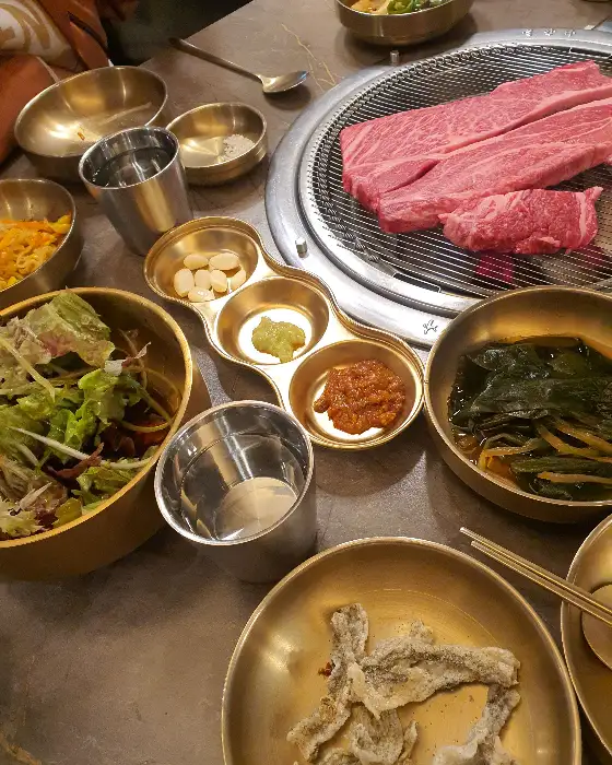 테이블에 차려진 고기와 곁들임 반찬들. 신선한 채소 샐러드, 나물 무침, 김치 등이 담긴 놋그릇들이 정갈하게 놓여 있다.