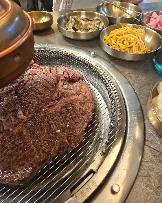 두툼한 소고기 덩어리가 숯불 위에서 지글지글 익어가고 있고, 주변으로 다양한 반찬이 담긴 놋그릇들이 놓여 있다.