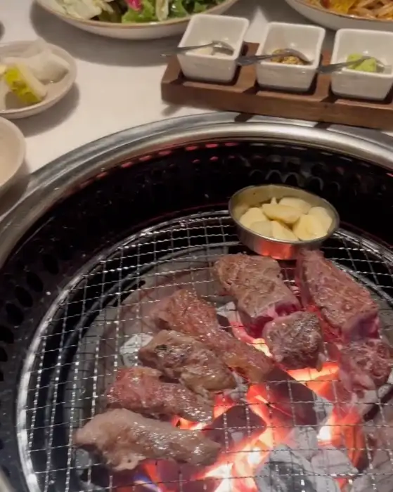 테이블에 놓인 숯불 구이 석쇠 위에 여러 조각의 소고기가 올려져 익고 있다. 주변에는 샐러드와 여러 종류의 소스가 담긴 작은 접시들이 놓여 있다.