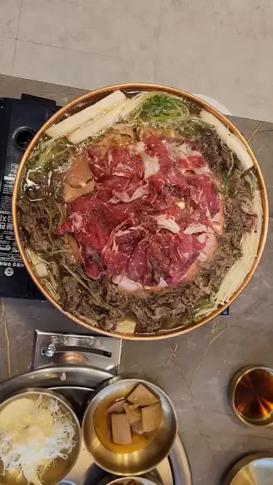 넓은 원형 불판 위에 숯이 담겨 있고, 그 위에 양념된 소고기와 채소가 올려져 끓여지고 있는 모습. 주변에는 놋그릇에 담긴 음식들이 보인다.