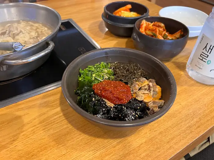 테이블 위에 꼬막비빔밥, 바지락칼국수, 반찬 등이 차려져 있다.