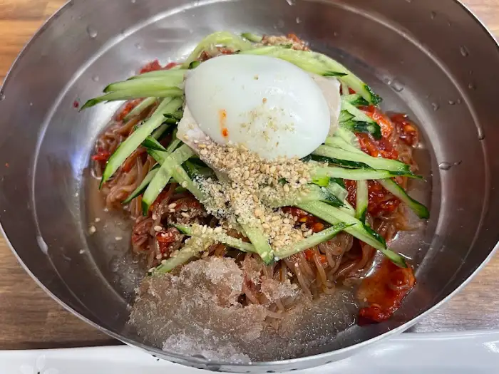 금속 쟁반에 담긴 비빔밀면의 모습입니다. 얇은 면 위에는 채 썬 오이, 반숙 달걀, 돼지고기 편이 올라가 있으며, 빨간 양념이 먹음직스럽습니다. 쟁반 가장자리에는 곱게 간 깨가 뿌려져 있습니다.