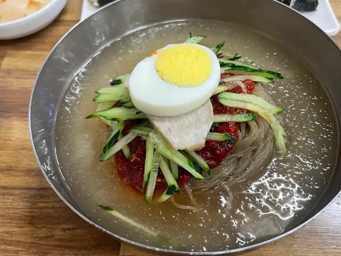 금속 쟁반에 담긴 물밀면의 모습입니다. 얇은 면 위에는 채 썬 오이, 반숙 달걀, 돼지고기 편이 올라가 있으며, 맑고 시원한 육수가 가득 담겨 있습니다. 쟁반 가장자리에는 곱게 간 깨가 뿌려져 있습니다.