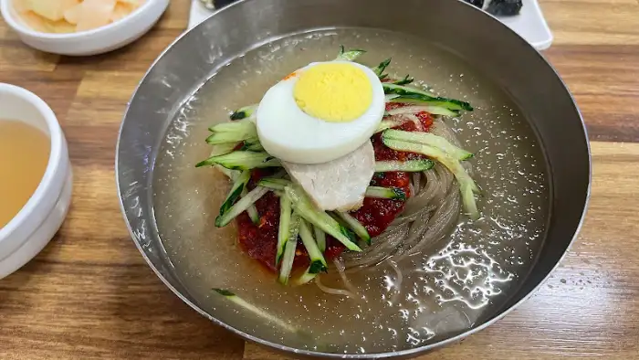 금속 쟁반에 담긴 물밀면의 모습입니다. 얇은 면 위에는 채 썬 오이, 반숙 달걀, 돼지고기 편이 올라가 있으며, 맑고 시원한 육수가 가득 담겨 있습니다. 쟁반 가장자리에는 곱게 간 깨가 뿌려져 있습니다.