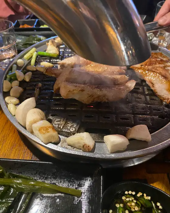 숯불 위에서 익어가고 있는 고기와 버섯, 파 등의 모습. 연기가 피어오르며 먹음직스럽다.